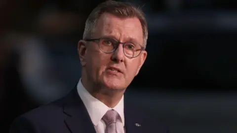A man is looking sideways, directly at the camera. He has short dark hair and is wearing rounded glasses. The man is dressed in a dark suit jacket with white shirt and light pink tie, which has a textured pattern. There is a small silver fish symbol attached to the lapel of his jacket. 