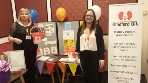 Bradford Teaching Hospitals Michaele and Sofia stand smiling next to a banner for their charity. Michaele is holding a donation bucket.