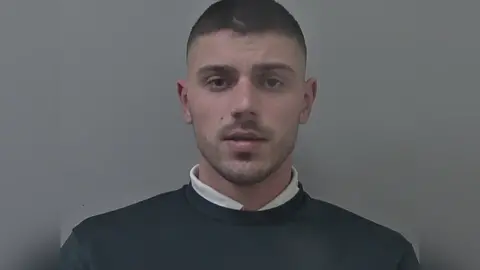 Man with dark short cropped hair and stubble. He is wearing a white shirt underneath a dark jumper.