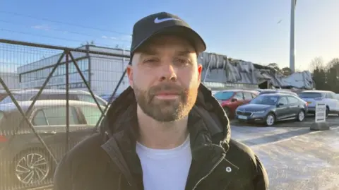Johnny Fusco, wearing black cap, with beard and black jacket in front of forecourt of cars.