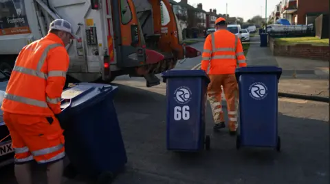 Getty Images Two Redcar and Cleveland Council workers collect bins. They are in a street of semi-detached houses. Both wear orange high-vis clothing and are wheeling three blue bins to the back of a refuse vehicle.