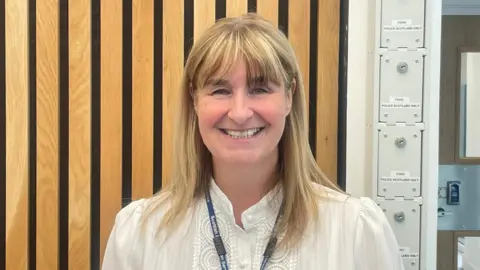 Pam Terry, a blonde-haired lady, stands against a wooden-slatted backdrop inside the prison. She is smiling and wears a white crocheted blouse and a Scottish Prison Service lanyard.