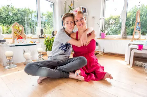 Alamy Alison Lapper cuddles her son Parys at their home in 2014. Alison is wearing a pink dress and a streak of her blonde hair is dyed pink. Parys is wearing a grey t-shirt and grey jeans. Artwork can be seen in the room behind them.