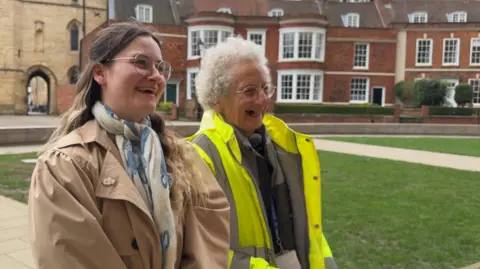 Two women laugh as they stand in a cathedral yard. The woman on the left is wearing a beige trench coat with a blue and white scarf and has long brown hair. Next to her is an older woman with white curly short hair, a yellow high-vis coat and lanyard. Behind them are houses and a medieval stone gatehouse.