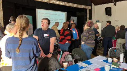 BBC/Richard Moss A group of people standing around talking