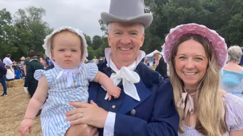 Baby, man and woman in Regency dress wearing bonnets and top hat at a Jane Austen celebration.