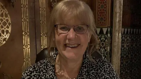 A woman with blonde hair and glasses and wearing a black and white spotty top, smiling at the camera with a patterned wall behind her.