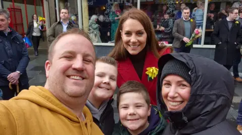 Gavin Halls' Gavin Halls, 41, who is wearing a mustard yellow hoodi, his son Ralfi, 12, who is wearing a black jumper and has a short pointed hair, Joseph, 8, which wearing a green puff jacket Are, and Aunty Hannah Jones. , 45. Princess Catherine smiles behind them. 