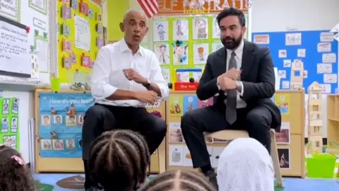 Barack Obama and Zohran Mamdani sit in front of a group of children singing