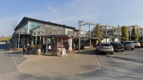 Google A picture of the front of the Darts Farm. There are cars parked outside the front of the shop with people walking around.