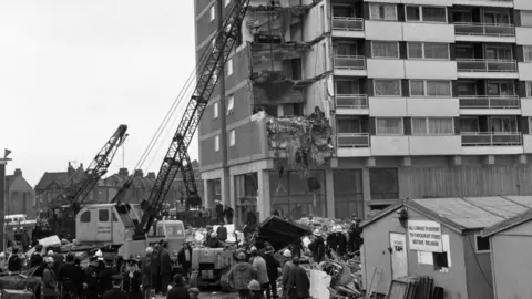 PA Media A black and white photo showing  multiple collapsed floors and walls on the corner of Ronan Point, with cranes stretching into the sky on the left. Crowds of people stand in front of a large pile rubble on the ground