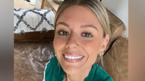 A smiling Hannah Viney taking a selfie in a room. She is sitting on a brown leather couch.