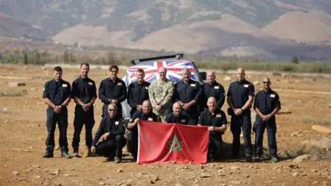 West Midlands Fire Service A group of men wearing black stand on sandy ground and hold a red flag with a green star on it. A car with a Union Jack flag on it is behind them. Mountains can be seen in the background.