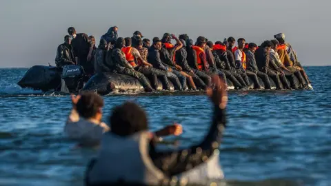 About thirty people are pictured on board a small black dinghy, in the Channel. Two people in the foreground are in the water and gesturing to the boat