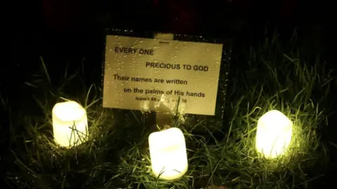 LDRS handout Three led candles on grass lit during a candlelight vigil at a cemetery to honour those buried in a mass grave with a yellow sign which reads: Every one precious to God: Their names are written on the palms of His hands. Isaiah 49 v 15 and 16 from the Bible 