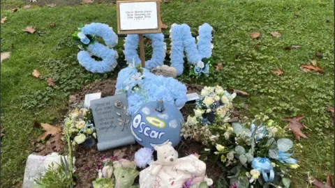 BBC A small grave, is surrounded by grass.  There is a blue wreath that says son in blue flowers and a blue pumpkin that says Oscar with a yellow halo above the name.