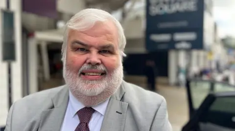 David Thomas, Conservative leader of Torbay Council, in a grey suit and a burgundy tie and standing on the pavement in Torbay with the sign for the Union Square shopping centre blurred in the background.