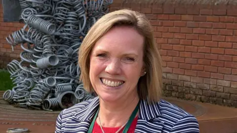 A woman wearing a blue and white striped jacket, with blonde hair in a long bob, smiling at the camera. Behind her, a brick wall and public artwork depicting a pile of metal springs.