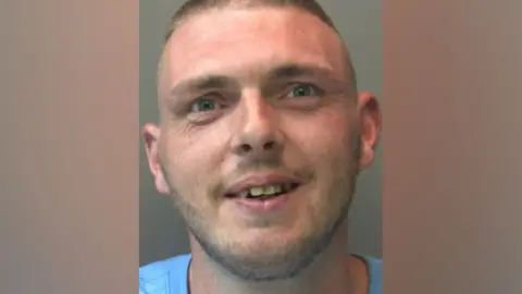 A police mugshot of Steven Stone, who has short, light-brown hair and stubble, and is smiling into the camera.