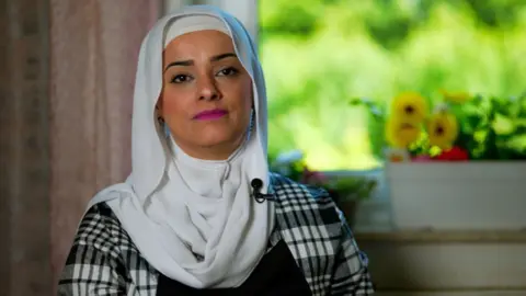 Headshot of a woman wearing a black and white checked top with a white headscarf. She is wearing pink lipstick. There is a pot with yellow flowers in the background.