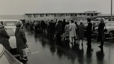 The Sainsbury Archive A black and white image of people queuing in a carpark in front of a Sainsbury's store in 1975