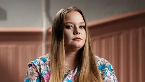 AB sits and sees the camera. Her long light brown hair and a floral dress.