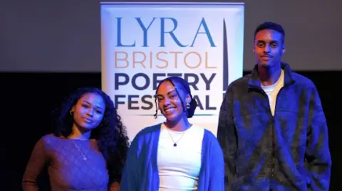 Sam Cavender Two young women and a young man stand are in a darkened room with a spotlight on them and a large poster behind saying 'Lyra Bristol Poetry Festival'. They are smiling to camera and wearing casual clothes