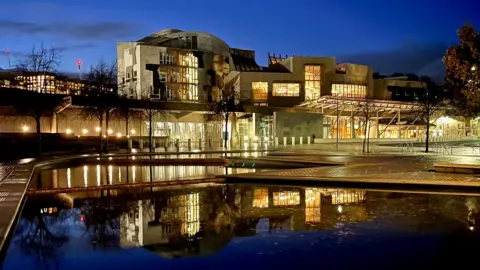 BBC A scenic photograph of the Scottish Parliament building in Edinburgh, reflected in the ponds outside its entrance, taken at night by the author of the article. The illuminated building glows yellow against the dark blue of the evening sky.
