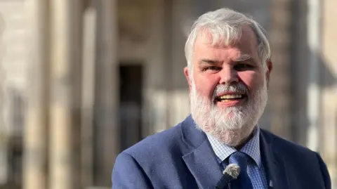 David Thomas in a blue check shirt and a blue tie and blue blazer with a blurred background of Torquay Town Hall.