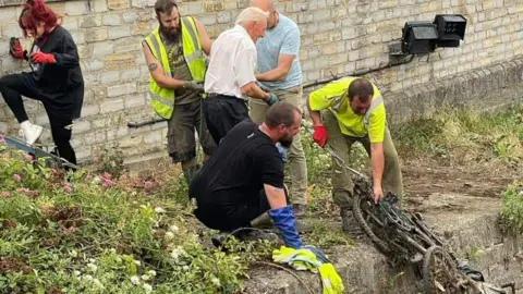 Terrence Pippin Volunteers get together to clean River Parrett. A group of people stand by the water's edge, some pulling debris from the river itself and others watching. Some of them are wearing high-vis jackets and protective gloves