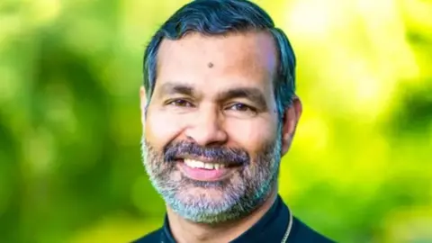 Church of England Right Reverend Dr John Perumbalath who has a grey beard smiles in front of a blurry green background