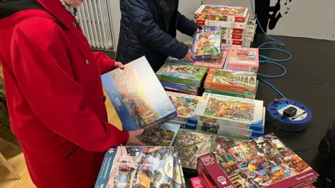 John Devine/BBC Two people seen from the neck down are looking through a pile of jigsaw puzzles on a low stage in a village hall.
