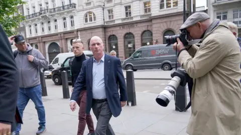 PA Media Ray Connolly with very short white hair, wearing a blue jacket and light blue shirt, with grey trousers, walking across a pavement pursued by press photographers. There is a road behind him.