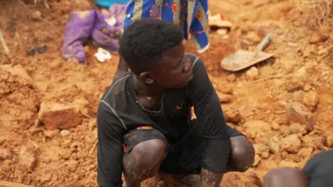 David Wilkins / BBC Komba Sesay, wearing black shorts and top is crouching in the light brown soil where he is mining.