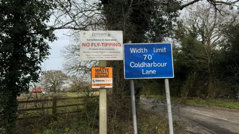 Three signs next to a country road in Egham. One says 'Runnymede Borough Council, Surveillance in Operation, NO FLY-TIPPING, fly-tippers will be prosecuted.' Another says 'WASTE CRIMINALS BEWARE, Selecta DNA IS USED IN THIS AREA.' The third days 'Width limit 7'0" Coldharbour Lane.'