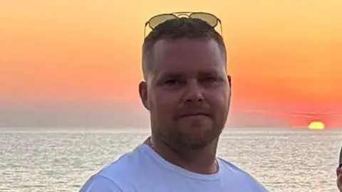 A man pictured wearing a t-shirt and sunglasses with a large body of water and a sunset in the background.
