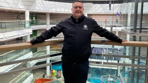 Andy is leaning back on a glass railing inside the centre, on top floors, and is smiling at the camera. He has a dark jacket and trousers on, as well as glass and has brown hair. Upper floors of the centre can be seen behind him.