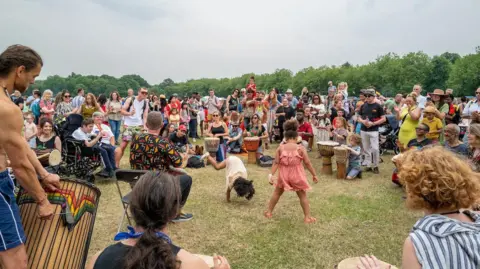 Africa Oye A large group of people are gathered in a circle on a large field with a row of trees in the background. Some people playing drums and dancing.