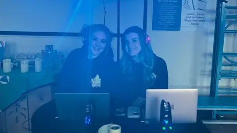 BBC / Ryan Dobney Jan and Ellie Hayes smile while sitting behind the mixing desk at the silent disco. They have computers in front of them and are wearing headphones.