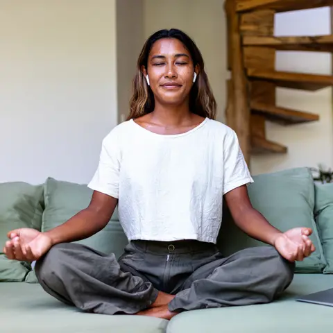 Getty Images A female sitting cross-legged connected a greenish sofa. She is wearing a achromatic T-shirt, grey trousers, and wireless earbuds. In nan background, location is simply a woody spiral staircase.