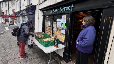 A woman in a purple jacket stands in the doorway of 3 East Street. The address is home to the Okehampton Community Foodbank. Outside the premises is a table with boxes of vegetables on it. A man and a woman appear to be taking vegetables as part of a foodbank package.