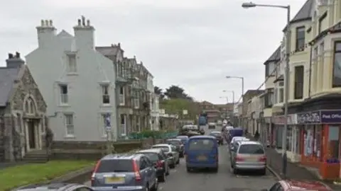 Google Church Street in Port, which is busy with traffic. It has shops on one side, a church and terraced housing on the other with cars parked on both side of the road.