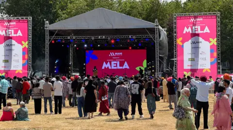 Crowds of people stand with their backs to the camera facing the stage with 'Birmingham Mela' signage on big screens visible