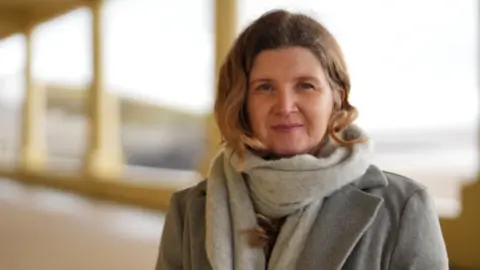 The BBC News Johanna Robinson looks towards the camera with a neutral expression. She wears a gray coat and a scarf and holds her blond hair behind her ears.