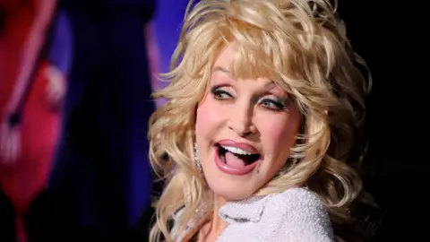Dolly Parton has her mouth open in a bright smile. She has voluminous blonde hair and is wearing a white top covered in rhinestones.