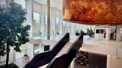 The entrance atrium of the building. It is a wide open space lined on one side by double-storey windows looking out over the trees. A large canopy covered in copper fish scales hangs over the reception desk and staircases lead up and down. There is a lecture room on the lower floor where an indoor tree is planted and glass cases display famous scientific instruments.