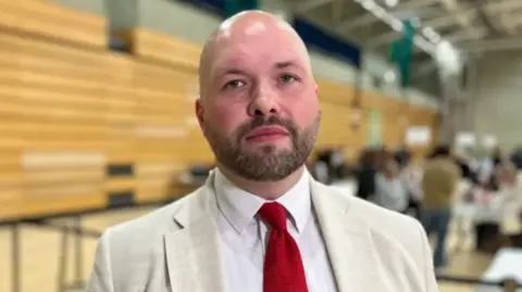 A person wearing a light‑coloured jacket and red tie stands indoors, facing the camera. The background is softly blurred, showing seating and activity inside a large hall.
