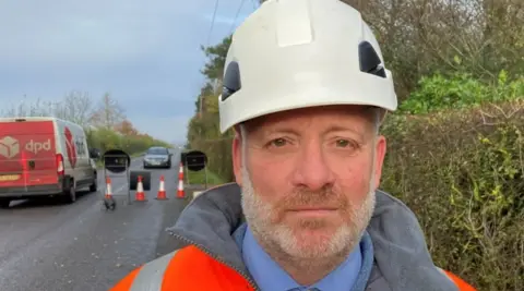 Robby West/BBC Darren Clark wearing a white hard hat with a road with cones behind him and a DPD van passing