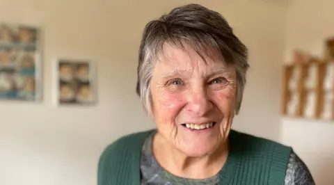 A picture of Margaret Gosley, wearing a green cardigan and grey jumper, photographed in her house smiling 