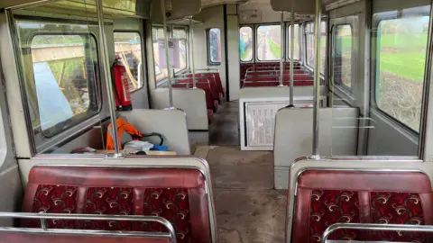 The interior of the railbus. The bus seats have been replaced with new burgundy seats which appear to be made of a mixture of leather and fabric. The bus is empty and has a fire extinguisher hanging on the right wall.